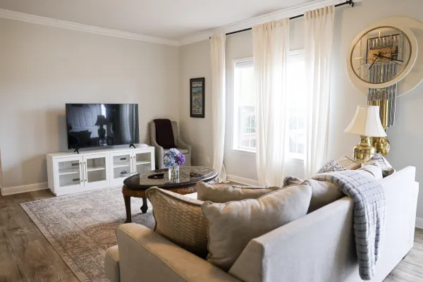 Bright living room with beige sofa, round wooden coffee table, TV on white stand, sheer curtains, and decorative wall clock.