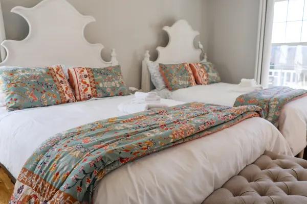 Two white beds with floral-patterned teal bedding and pillows in a bright, cozy bedroom with natural light.