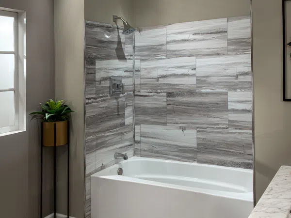 Modern bathroom with white bathtub, gray stone tile walls, a window, and a potted plant in a gold and black stand.