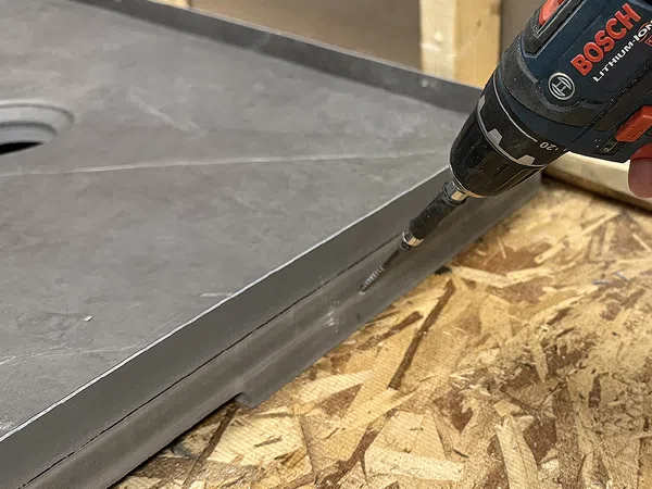Person using drill to assemble gray shower pan on wooden surface.