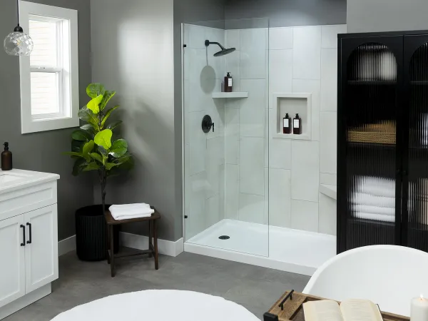 Modern bathroom with glass shower, white tub, black cabinet, green plant, and wooden stool with towels