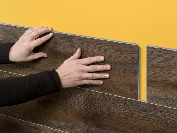 Hands installing dark wood laminate panels on a yellow wall, showing wall paneling process and texture.