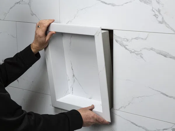 Hands installing a white marble-patterned recessed shower niche into a tiled bathroom wall with matching marble tiles.