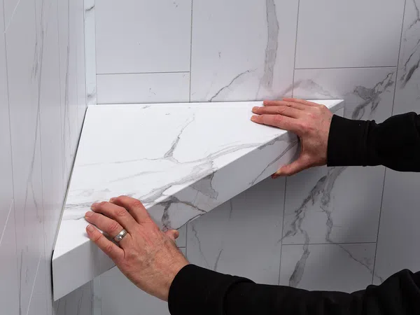 Person installing a white marble-patterned triangular shelf corner on matching tiled wall in a bathroom.