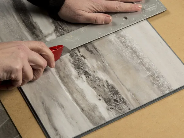 Person cutting a marble-patterned tile with a red utility knife using a metal ruler on a wooden surface