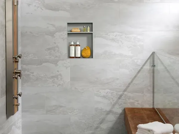 Modern bathroom shower area with gray marble tiles, rain showerhead, wooden bench, and glass partition.