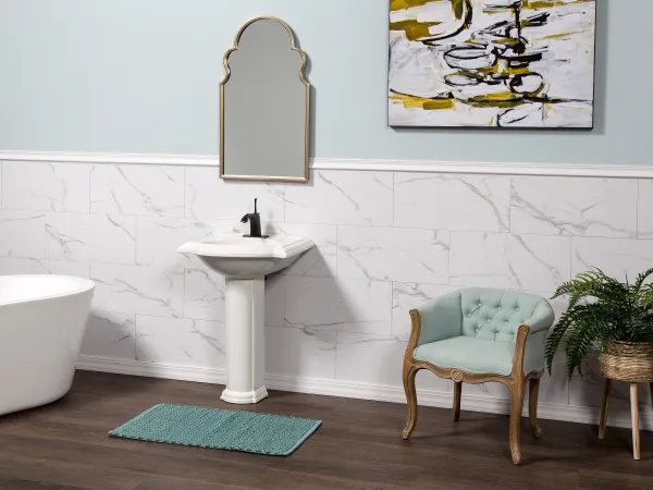 Modern bathroom with white pedestal sink, arched mirror, tub, teal chair, plant, and abstract wall art.