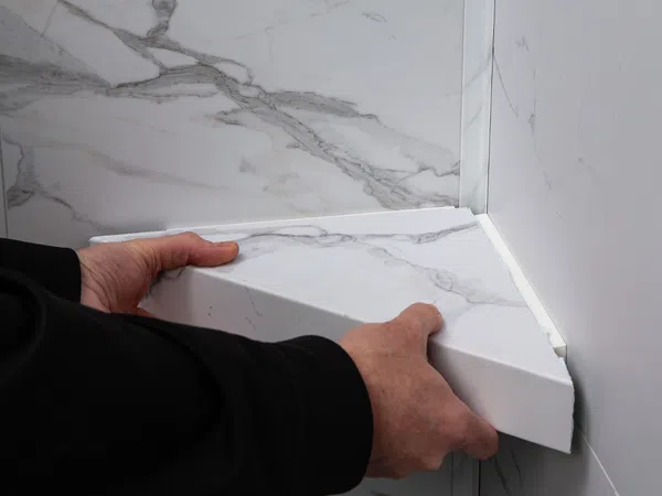 Hands installing a white marble-patterned corner shelf in a bathroom or kitchen wall corner.