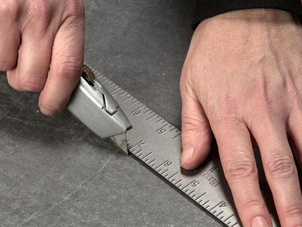 Person cutting a straight line on a gray surface using a metal ruler and utility knife.