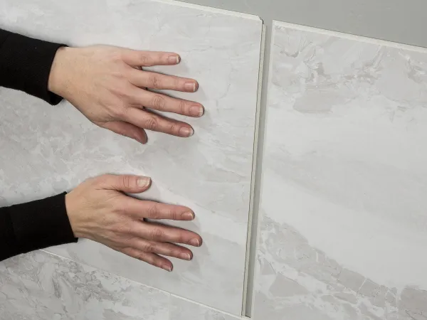 Hands installing large light gray marble wall tiles with subtle veining on a vertical surface indoors.