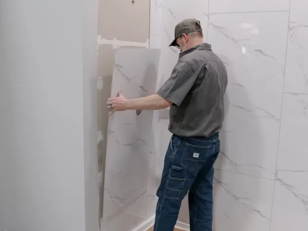 Worker installing large marble-patterned wall panels in a bathroom under construction.