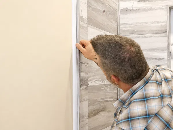 Man installing wallpaper on a tiled bathroom wall near a window wearing a plaid shirt.
