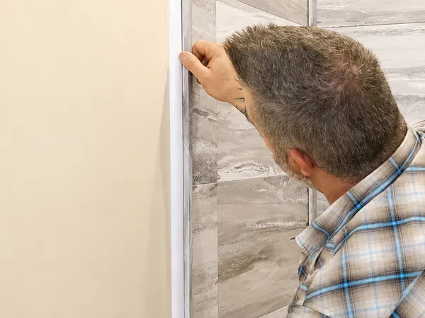 Man installing trim on wall corner next to tiled bathroom area with natural light window
