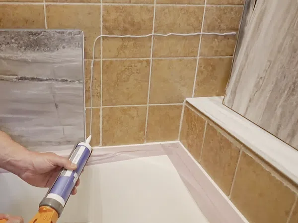 Person applying white caulk with a caulking gun around a bathtub edge next to beige tiled walls