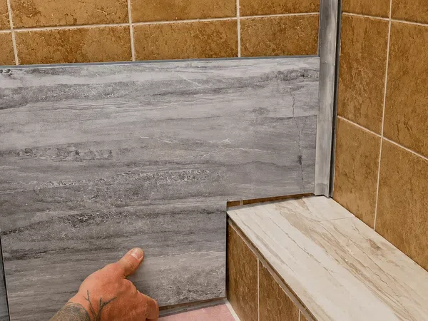 Hands installing gray marble-look tile on a step in a bathroom with brown wall tiles.