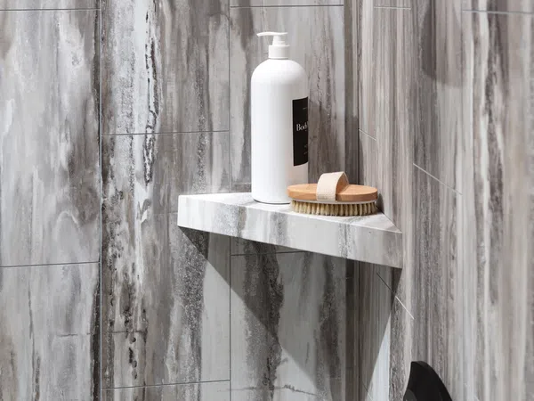 Shower corner with gray marble tiles, a white lotion bottle, and a wooden brush on a marble shelf.
