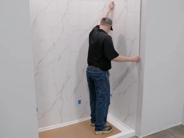 Man installing large marble-look wall panels in a shower space with protective floor covering in a bathroom.