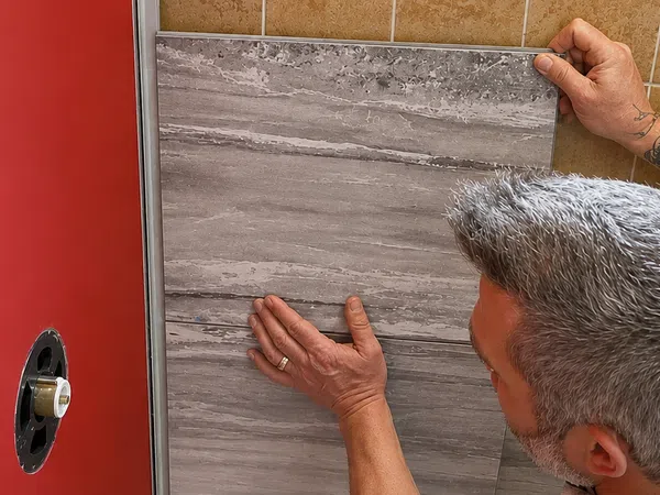 Man with tattooed arm installing large gray tile on bathroom wall next to red door.