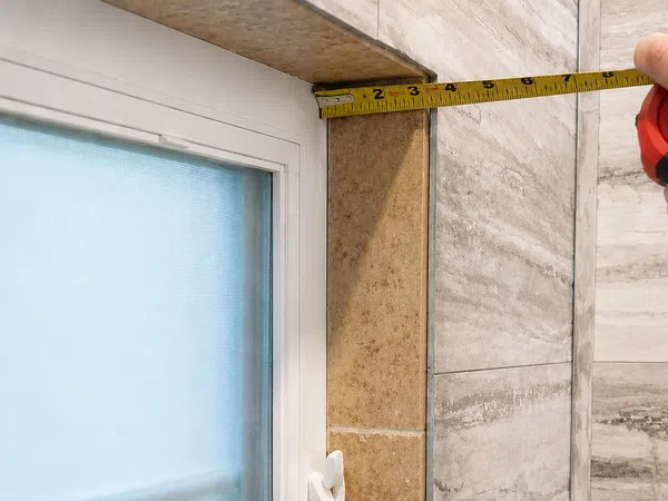 Hand holding tape measure to check length of window frame against tiled wall in home interior.