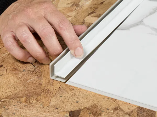 Hand installing white trim or tile edging on wooden surface next to a white tile with marble pattern.