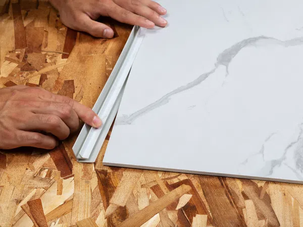 Hands measuring a white marble tile edge on a wooden surface using a plastic straightedge tool for precision.