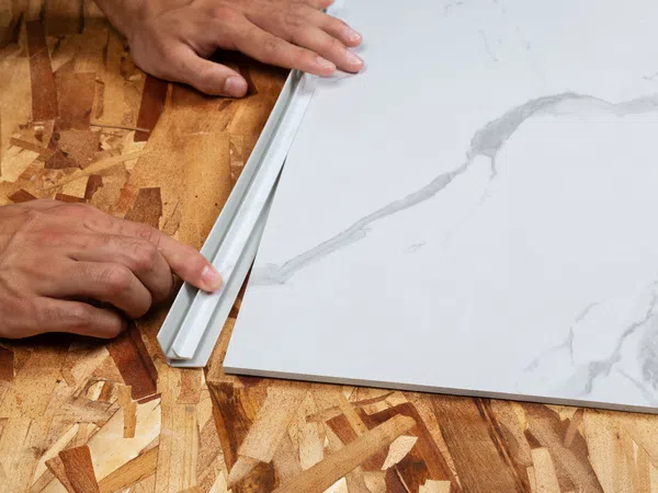 Hands installing white marble-patterned tile with plastic trim on plywood subfloor surface.