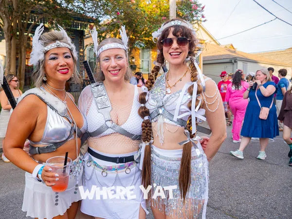 Three women in futuristic silver festival costumes posing outdoors at a sunny street event with a festive crowd.
