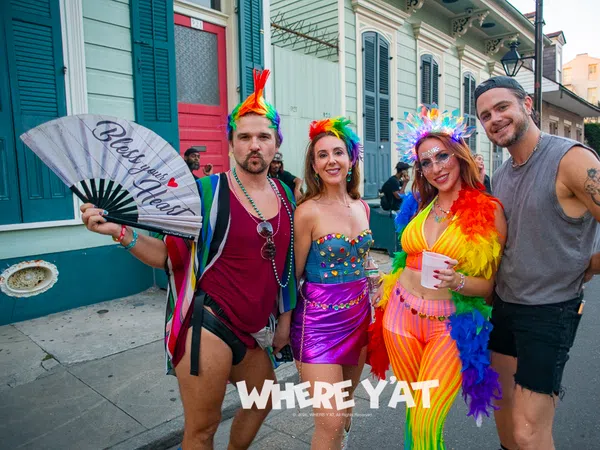 Four colorful, lively people in festive rainbow outfits posing on a New Orleans street during a celebration.