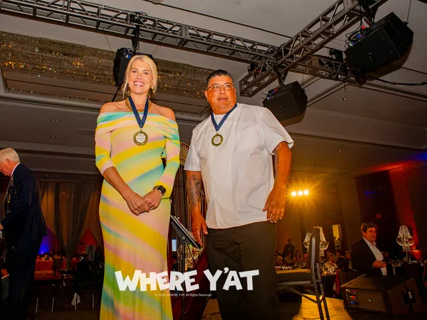 Two award recipients wearing medals stand on stage at a formal event with audience in background.