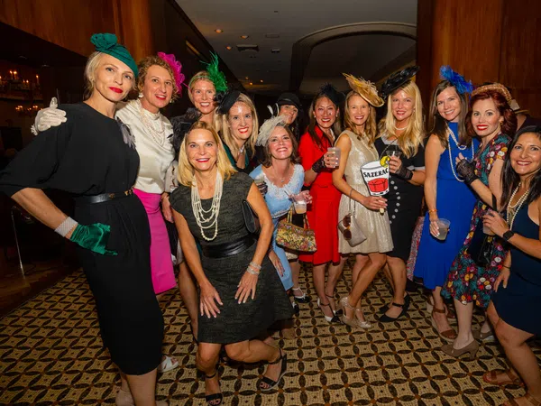 Group of women dressed in vintage-style dresses and hats posing indoors for a lively social event photo.