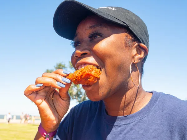 Smiling woman wearing a black cap and hoop earrings eating fried chicken outdoors on a sunny day