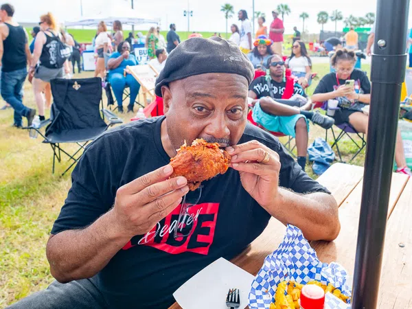 Man in black hat eating fried chicken at outdoor event with people and tents in the background
