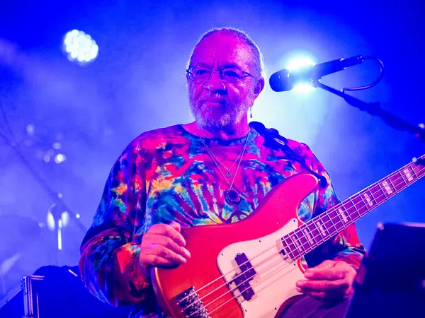 Musician wearing a colorful tie-dye shirt playing red electric bass guitar on stage with blue lighting.