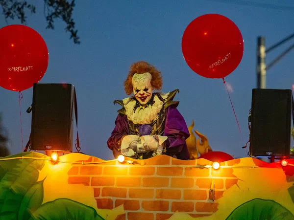 Costumed clown with red hair and white face paint holding red balloons at night during parade float event.