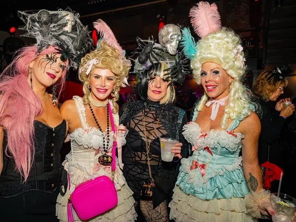 Four women in elaborate Halloween costumes with wigs and accessories at a festive party indoors.