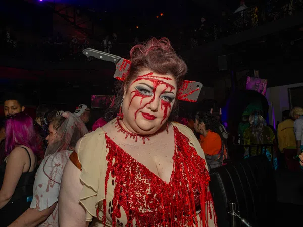 Woman in bloody costume with fake cleaver through head at a Halloween party with crowd in background