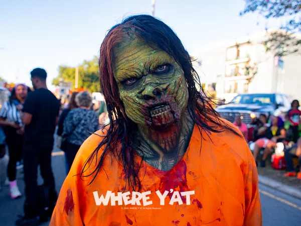 Person in detailed zombie makeup wearing a blood-stained orange shirt with 'WHERE Y'AT' text, outdoors at event