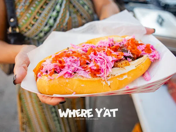 Close-up of a hand holding a hot dog with pink slaw and red sauce in a paper tray, with overlaid text reading WHERE Y'AT.