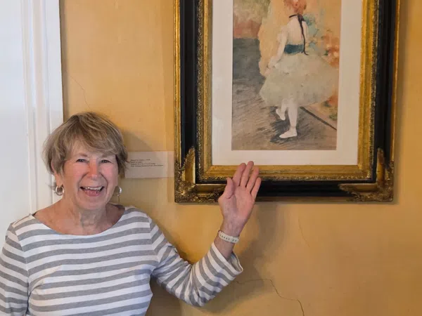 Smiling elderly woman pointing at framed impressionist painting of ballerina in museum setting