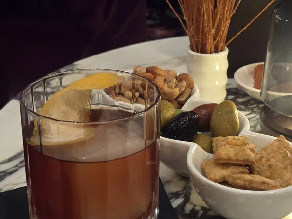 Old fashioned cocktail with lemon peel served with assorted nuts, olives, crackers, and breadsticks on marble table.