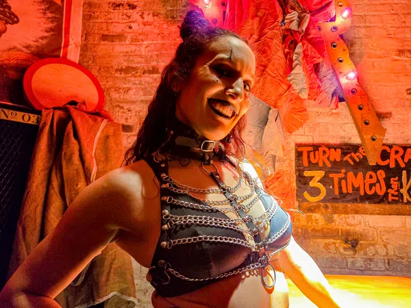 Woman in black leather harness and chains smiles against a brick wall with Halloween decor and overlaid text sign.