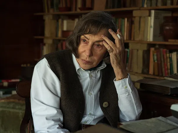 Elderly woman with brown hair sitting contemplatively in a vintage study room filled with books and a typewriter.