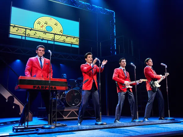 Four male performers in red jackets play instruments and sing on stage with musical notes backdrop in blue lighting.