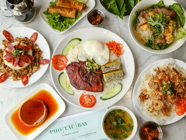 Overhead view of Vietnamese dishes including grilled pork with egg, spring rolls, noodle soup, shrimp skewers, and caramel flan on a marble table.
