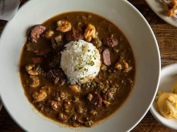 Bowl of shrimp and sausage gumbo with white rice and herbs on top, served on a wooden table with side dishes.