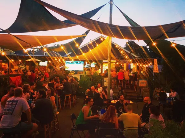 Outdoor evening bar scene with string lights, shaded canopies, and groups of people socializing under warm lighting.