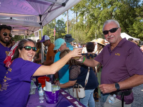 People enjoying an outdoor event exchanging drinks under purple tents on a sunny day with trees in the background