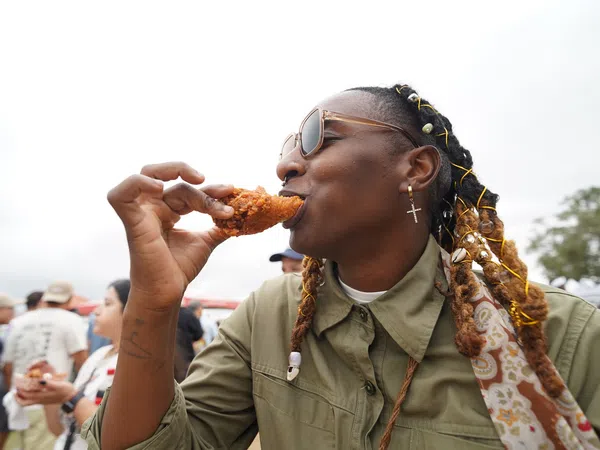 Person with braided hair and sunglasses enjoying crispy fried chicken outdoors at a casual event.