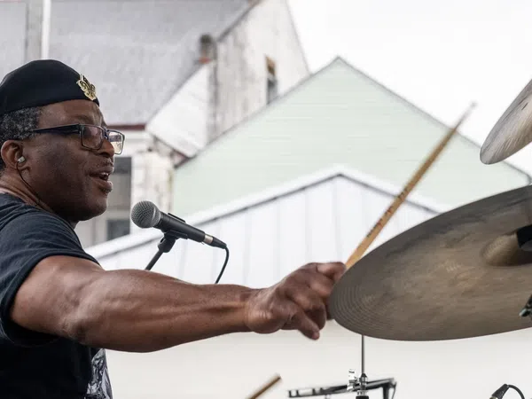 Man wearing a beret playing drums energetically at an outdoor performance with houses in the background