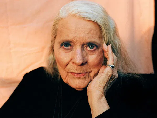 Elderly woman with silver hair, wearing black, resting her face on her hand against a soft peach background.